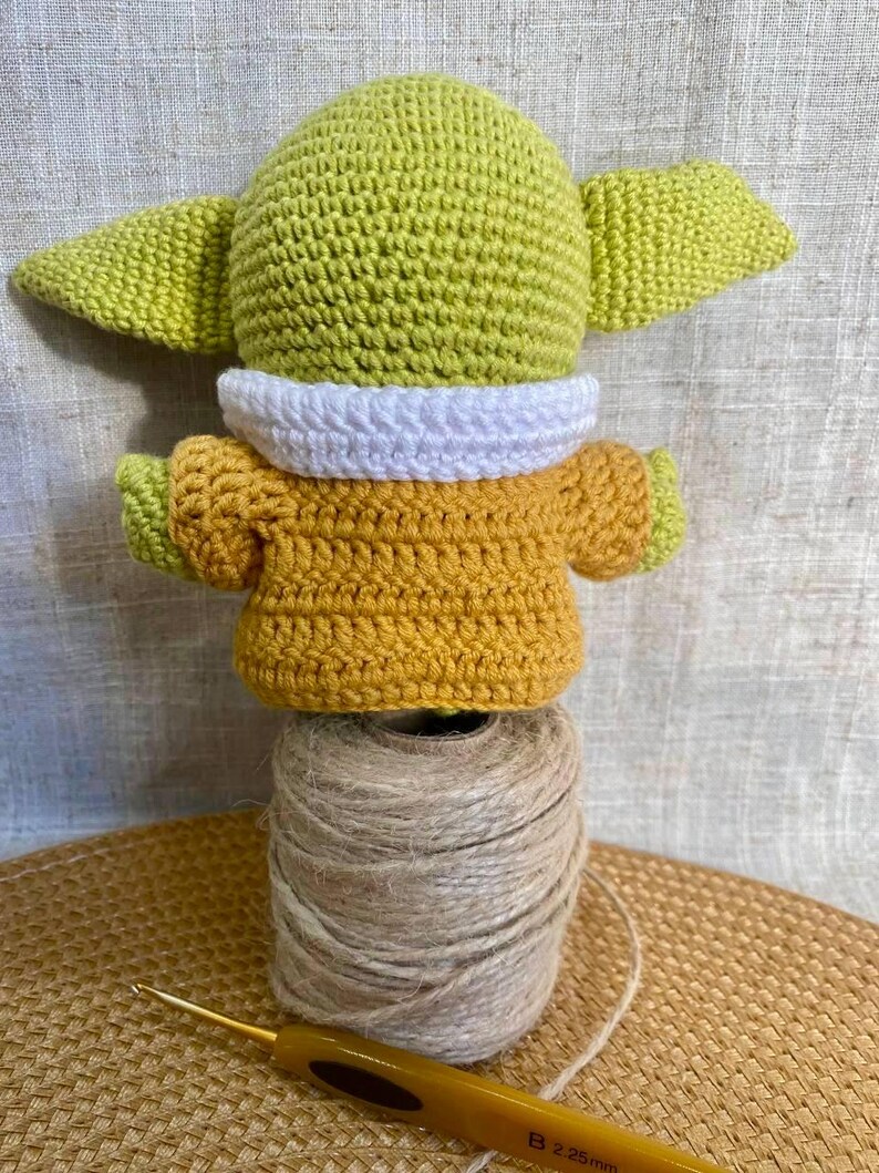 May include: A handmade crochet figure of a character with large ears, wearing a yellow and white outfit. The figure is perched atop a spool of twine, with a crochet hook in the foreground. The figure is approximately 4 inches tall.