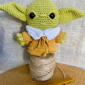 May include: A handmade crochet doll of a character with large ears, wearing a yellow and white outfit. The doll is displayed on a spool of twine with a yellow crochet hook. The doll's head and ears are light green, and the eyes are black.