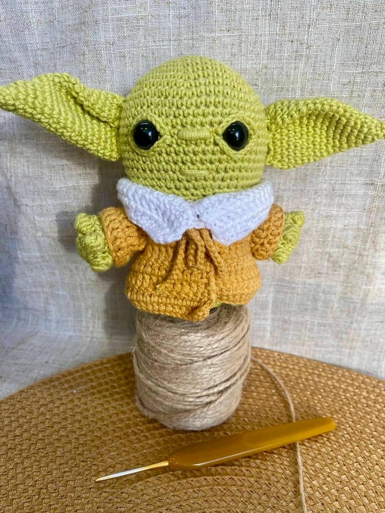 May include: A handmade crochet doll of a character with large ears, wearing a yellow and white outfit. The doll is displayed on a spool of twine with a yellow crochet hook. The doll's head and ears are light green, and the eyes are black.
