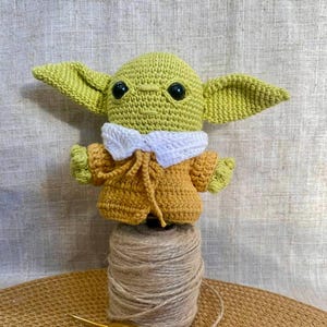 May include: A handmade crochet figure of a character with large ears, green skin, and black eyes. The figure wears a yellow coat with a white collar. It is perched on a spool of twine, with a crochet hook nearby.