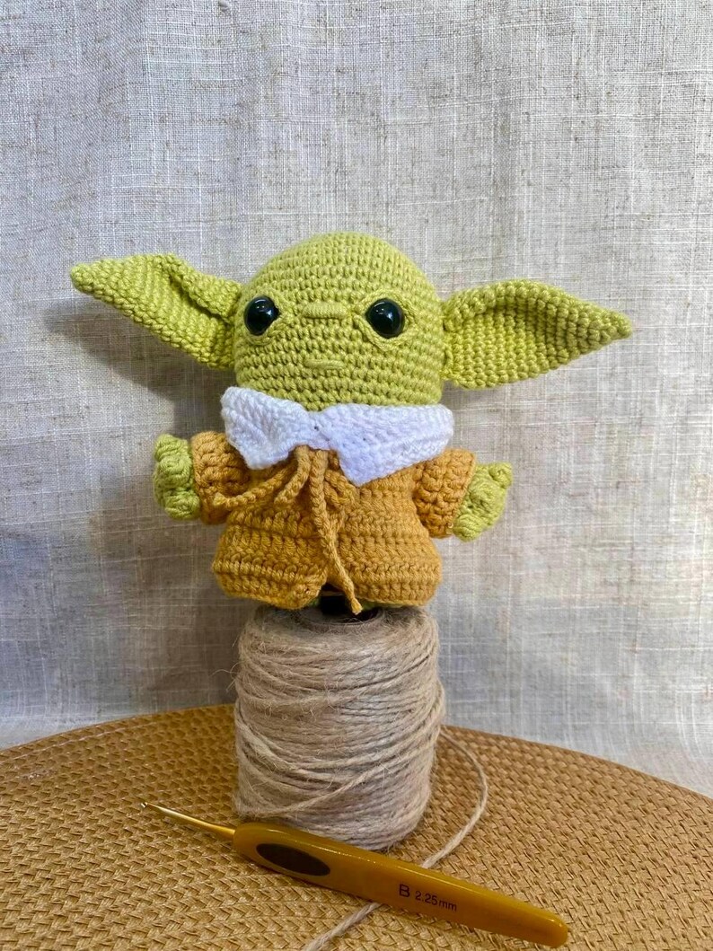 May include: A handmade crochet figure of a character with large ears, green skin, and black eyes. The figure wears a yellow coat with a white collar. It is perched on a spool of twine, with a crochet hook nearby.