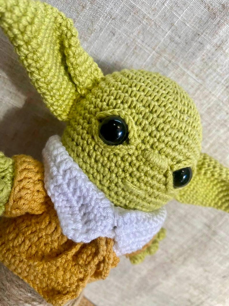 May include: Crocheted green character with large black eyes, long ears, and a white collar. The character is wearing a yellow and green outfit. The handmade doll is a popular character from a science fiction series.