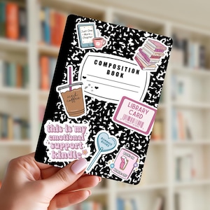 May include: A black and white composition book with various stickers. Stickers include a coffee cup, a stack of books, a library card, and the text "this is my emotional support Kindle". The book is held in a hand.