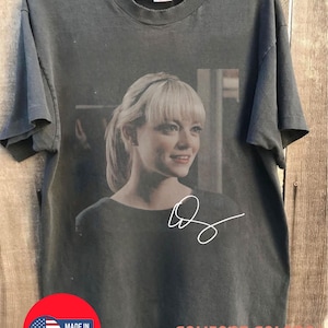 May include: A Comfort Colors pepper-colored t-shirt featuring a portrait of a woman with blonde hair and a signature. The shirt has a round neckline and short sleeves. A red circle with "Made in USA" is visible.
