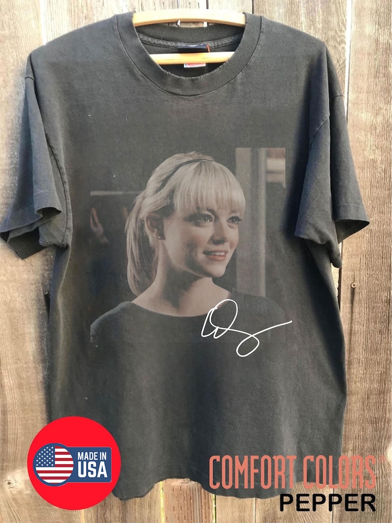 May include: A Comfort Colors pepper-colored t-shirt featuring a portrait of a woman with blonde hair and a signature. The shirt has a round neckline and short sleeves. A red circle with "Made in USA" is visible.