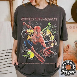 May include: Dark gray t-shirt featuring a Spider-Man graphic. The design includes Spider-Man in a dynamic pose, the text "SPIDER-MAN", and the phrase "WITH GREAT POWER COMES GREAT RESPONSIBILITY". The shirt is from Comfort Colors.