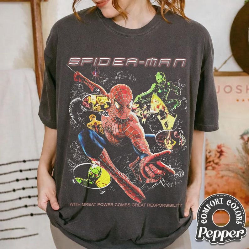 May include: Dark gray t-shirt featuring a Spider-Man graphic. The design includes Spider-Man in a dynamic pose, the text "SPIDER-MAN", and the phrase "WITH GREAT POWER COMES GREAT RESPONSIBILITY". The shirt is from Comfort Colors.