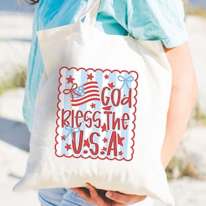 May include: A cream-colored tote bag with a patriotic design. The design features the American flag, stars, and the text "God Bless The U.S.A." in red, white, and blue. The bag is held by a person wearing a light blue shirt.