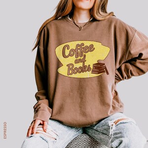 May include: Brown sweatshirt with the text "Coffee and Books" in a yellow oval. The design includes a stack of books and a coffee cup. The sweatshirt is a neutral color and has a relaxed fit.