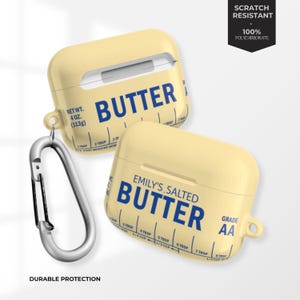 Puede incluir: Dos fundas amarillas para AirPods con un diseño temático de mantequilla. Las fundas presentan la palabra "BUTTER" en azul, junto con marcas de medición. Una funda tiene impreso "EMILY'S SALTED BUTTER". Las fundas son resistentes a los arañazos y están hechas de 100% policarbonato.