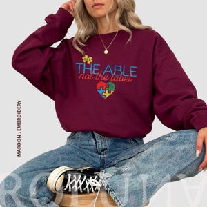 Puede incluir: Sudadera granate con el texto bordado "SEE THE ABLE not the label" y un dise&ntilde;o de coraz&oacute;n de rompecabezas en azul, verde, amarillo y rojo. La sudadera es de cuello redondo.