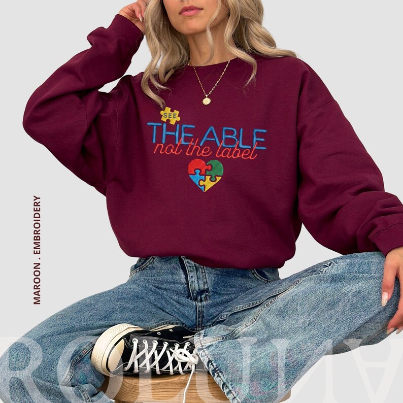 Puede incluir: Sudadera granate con el texto bordado "SEE THE ABLE not the label" y un dise&ntilde;o de coraz&oacute;n de rompecabezas en azul, verde, amarillo y rojo. La sudadera es de cuello redondo.
