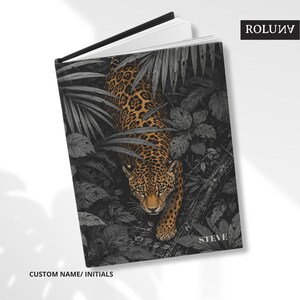 May include: Open journal with a cover depicting a leopard in a jungle scene. The leopard's coat is tan and brown with spots, set against a grey-scale jungle. The cover includes the name "STEVE" and the text "CUSTOM NAME/ INITIALS". The journal has a black spine.
