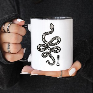 Könnte beinhalten: Eine weiße Keramik-Tasse mit schwarzem Henkel und dem Text "But First, Coffee" in schwarzer, handschriftlicher Schrift. Die Tasse steht auf einer weißen Oberfläche.