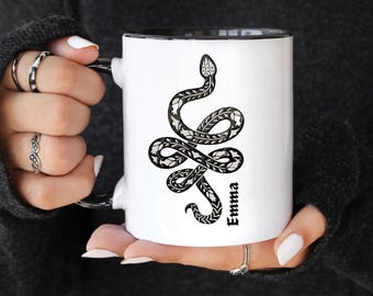 May include: A white ceramic mug with a black interior and handle, featuring a black graphic illustration of a coiled snake with intricate floral patterns on its body and the name Emma printed vertically in black text next to the snake.