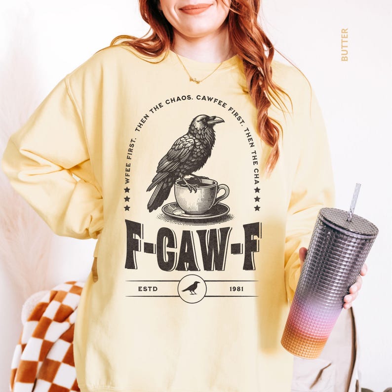 May include: A light yellow sweatshirt with a black graphic of a raven perched on a teacup, with the text "F-CAW-F" and "CAWFEE FIRST. THEN THE CHAOS." A gradient tumbler is held in the hand.