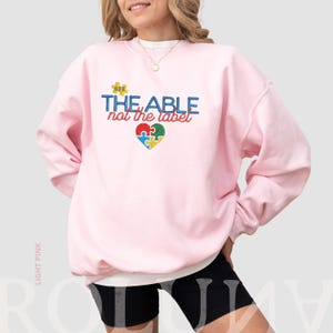 Puede incluir: Sudadera rosa claro con el texto "SEE THE ABLE not the label" en azul, rojo y amarillo. Un dise&ntilde;o de rompecabezas en forma de coraz&oacute;n en el centro presenta piezas azules, verdes, rojas y amarillas. La sudadera es de cuello redondo.
