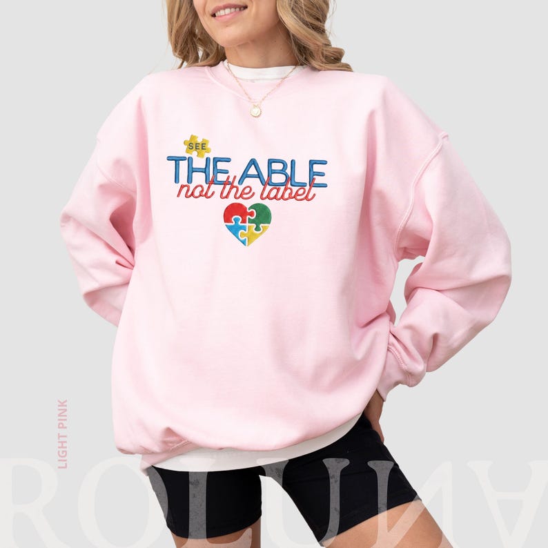 Puede incluir: Sudadera rosa claro con el texto "SEE THE ABLE not the label" en azul, rojo y amarillo. Un dise&ntilde;o de rompecabezas en forma de coraz&oacute;n en el centro presenta piezas azules, verdes, rojas y amarillas. La sudadera es de cuello redondo.