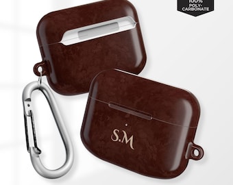 May include: Two brown AirPod cases with a textured finish, one with the initials "S.M." in gold. A silver carabiner clip is attached to one case. The cases are labelled "Scratch Resistant" and "Durable Protection."