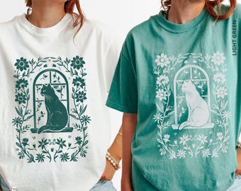 May include: White and light green t-shirts featuring a cat silhouette in a window, surrounded by floral designs. The white shirt has a dark green graphic, while the light green shirt has a white graphic. The shirts are casual and comfortable.