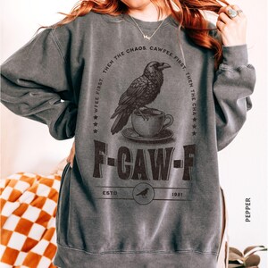May include: Dark gray sweatshirt with a graphic of a raven perched on a teacup. The text reads "WFEE FIRST, THEN THE CHAOS. CAWFEE FIRST. THEN THE CHA" and "F-CAW-F ESTD 1981". The sweatshirt is labeled "PEPPER".