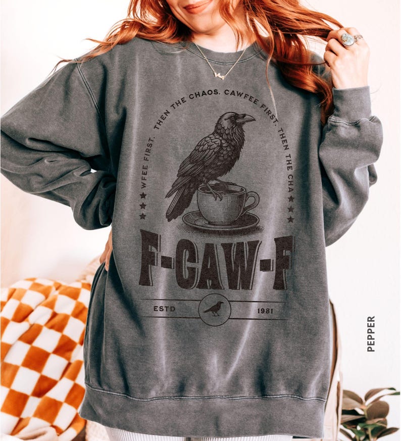May include: Dark gray sweatshirt with a graphic of a raven perched on a teacup. The text reads "WFEE FIRST, THEN THE CHAOS. CAWFEE FIRST. THEN THE CHA" and "F-CAW-F ESTD 1981". The sweatshirt is labeled "PEPPER".