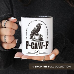 May include: White ceramic mug with a black interior and handle. The mug features a black and white illustration of a raven perched above a teacup, with the text "F-CAW-F" and "CAWFEE FIRST. THEN THE CHAOS."