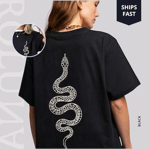 Puede incluir: Camiseta negra con un diseño de serpiente blanca en la espalda. La ilustración de la serpiente es detallada y estilizada. La camiseta es de estilo cuello redondo clásico. La palabra "BLACK" está impresa verticalmente en el lado derecho de la camiseta.