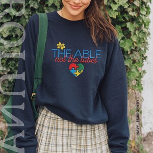 Puede incluir: Sudadera azul marino con la frase bordada "SEE THE ABLE not the label" y un dise&ntilde;o de coraz&oacute;n con piezas de rompecabezas. La sudadera es de cuello redondo.