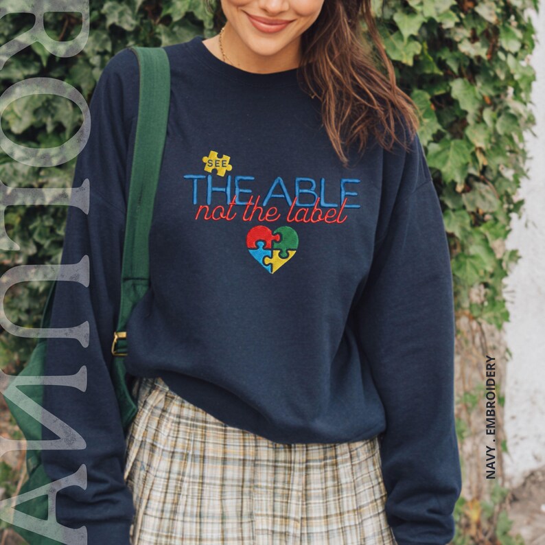 Puede incluir: Sudadera azul marino con la frase bordada "SEE THE ABLE not the label" y un dise&ntilde;o de coraz&oacute;n con piezas de rompecabezas. La sudadera es de cuello redondo.