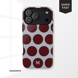 Puede incluir: Una funda de teléfono con un patrón de lunares granates grandes sobre un fondo gris claro. La funda tiene un módulo de cámara negro y la letra "M" en la parte inferior. Las palabras "MAGSAFE®" están en la parte inferior izquierda y "SHIPS FAST" en una pancarta negra.