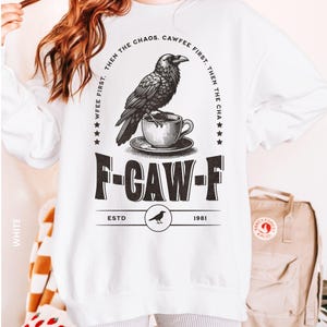 May include: White sweatshirt with a black graphic of a raven perched on a teacup. The text reads "WFEE FIRST. THEN THE CHAOS. CAWFEE FIRST. THEN THE CHA" and "F-CAW-F ESTD 1981". A casual, long-sleeved top.