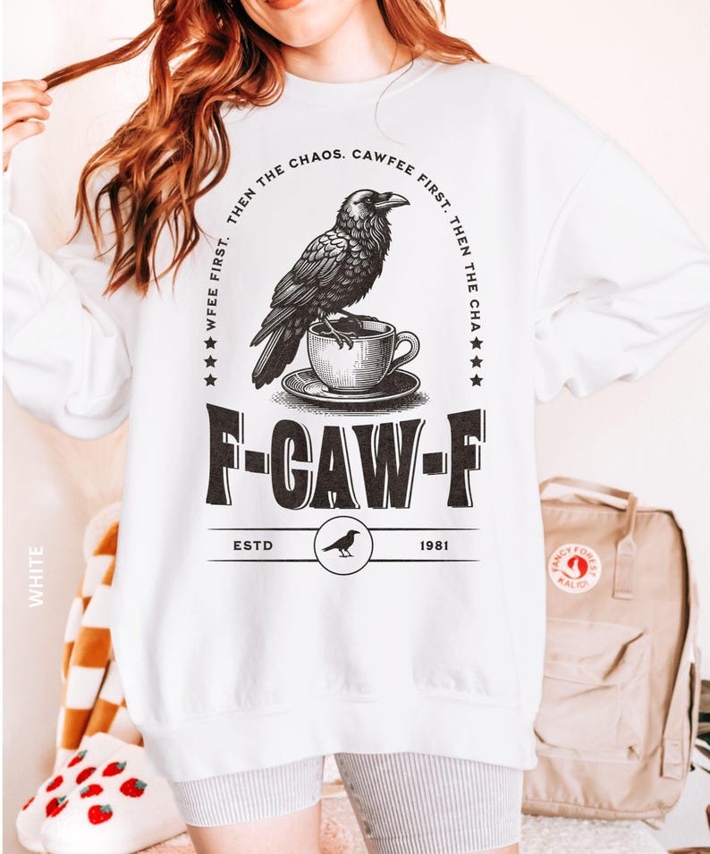 May include: White sweatshirt with a black graphic of a raven perched on a teacup. The text reads "WFEE FIRST. THEN THE CHAOS. CAWFEE FIRST. THEN THE CHA" and "F-CAW-F ESTD 1981". A casual, long-sleeved top.