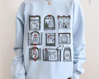 May include: Light blue sweatshirt featuring a whimsical design of ghosts in Christmas-themed windows. Each window showcases a different festive scene, including Christmas trees, fireplaces, and snowmen. The sweatshirt is a comfortable, casual style.