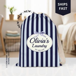 May include: A navy blue and white striped laundry bag with a drawstring closure. The bag features an oval label with the text "Olivia's Laundry" and a phone number. A light beige drawstring and a "Ships Fast" label are also visible.