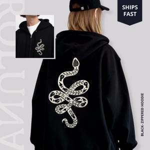 Könnte beinhalten: Schwarzer Kapuzenpullover mit Reißverschluss und detailliertem weißen Schlangenmuster auf dem Rücken und einer kleineren Version auf der Vorderseite. Der Hoodie hat einen Frontreißverschluss und eine Kapuze. Der Text "BLACK- ZIPPERED HOODIE" ist gedruckt.