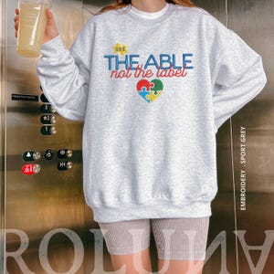 Puede incluir: Una sudadera gris claro con el texto bordado "SEE THE ABLE not the label" en azul, rojo y amarillo. Un coraz&oacute;n hecho de piezas de rompecabezas est&aacute; debajo del texto. La sudadera es gris deportivo.