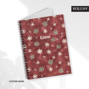 Può includere: Un quaderno a spirale con una copertina bordeaux ornata da motivi a fiocchi di neve bianchi e verdi. Il nome "Emma" è stampato in bianco. Le parole "CUSTOM NAME" sono stampate in basso. Il quaderno è aperto, rivelando pagine bianche a righe.