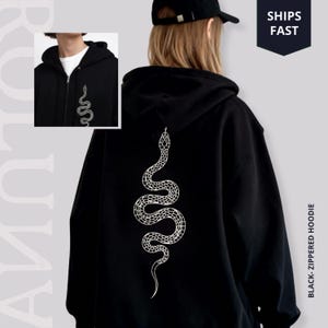 Può includere: Felpa con cappuccio nera con cerniera e design a serpente bianco sul retro e sul davanti. La felpa con cappuccio ha una cerniera completa e un cappuccio. L'immagine include anche il testo "SHIPS FAST" e "BLACK-ZIPPERED HOODIE".