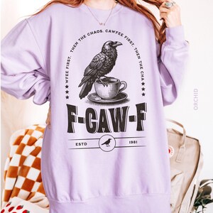 May include: A light purple sweatshirt with a graphic featuring a raven perched on a teacup, with the text "F-CAW-F" and "CAWFEE FIRST. THEN THE CHAOS." The sweatshirt has a crew neck and long sleeves. The text "ESTD 1981" is also visible.