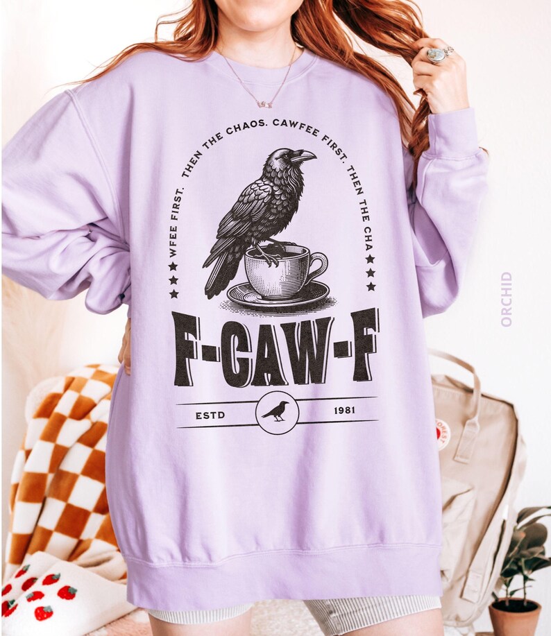 May include: A light purple sweatshirt with a graphic featuring a raven perched on a teacup, with the text "F-CAW-F" and "CAWFEE FIRST. THEN THE CHAOS." The sweatshirt has a crew neck and long sleeves. The text "ESTD 1981" is also visible.