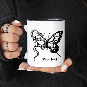 May include: White ceramic mug with a black interior and handle. The mug features a black and white graphic of a snake intertwined with a butterfly, with the text "Your text" below the graphic.