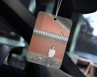 May include: Rectangular car air freshener with a photograph of a person standing in front of a red brick wall. The air freshener is hanging from a string, with a blurred car interior in the background.
