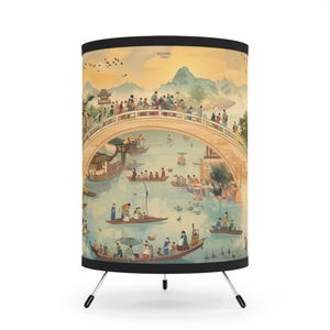 Peut inclure: Lampe de table tripode avec une illustration détaillée d'une scène chinoise traditionnelle. L'œuvre représente un pont, des bateaux, des personnes et des montagnes dans un paysage serein et coloré. La lampe a une bordure noire et un fond blanc.