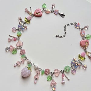 May include: A delicate necklace featuring a variety of pastel-colored beads and charms. The necklace includes pink heart-shaped pendants, flower and leaf charms, and a cluster of light purple beads. The chain is silver-toned, and the overall design is whimsical.