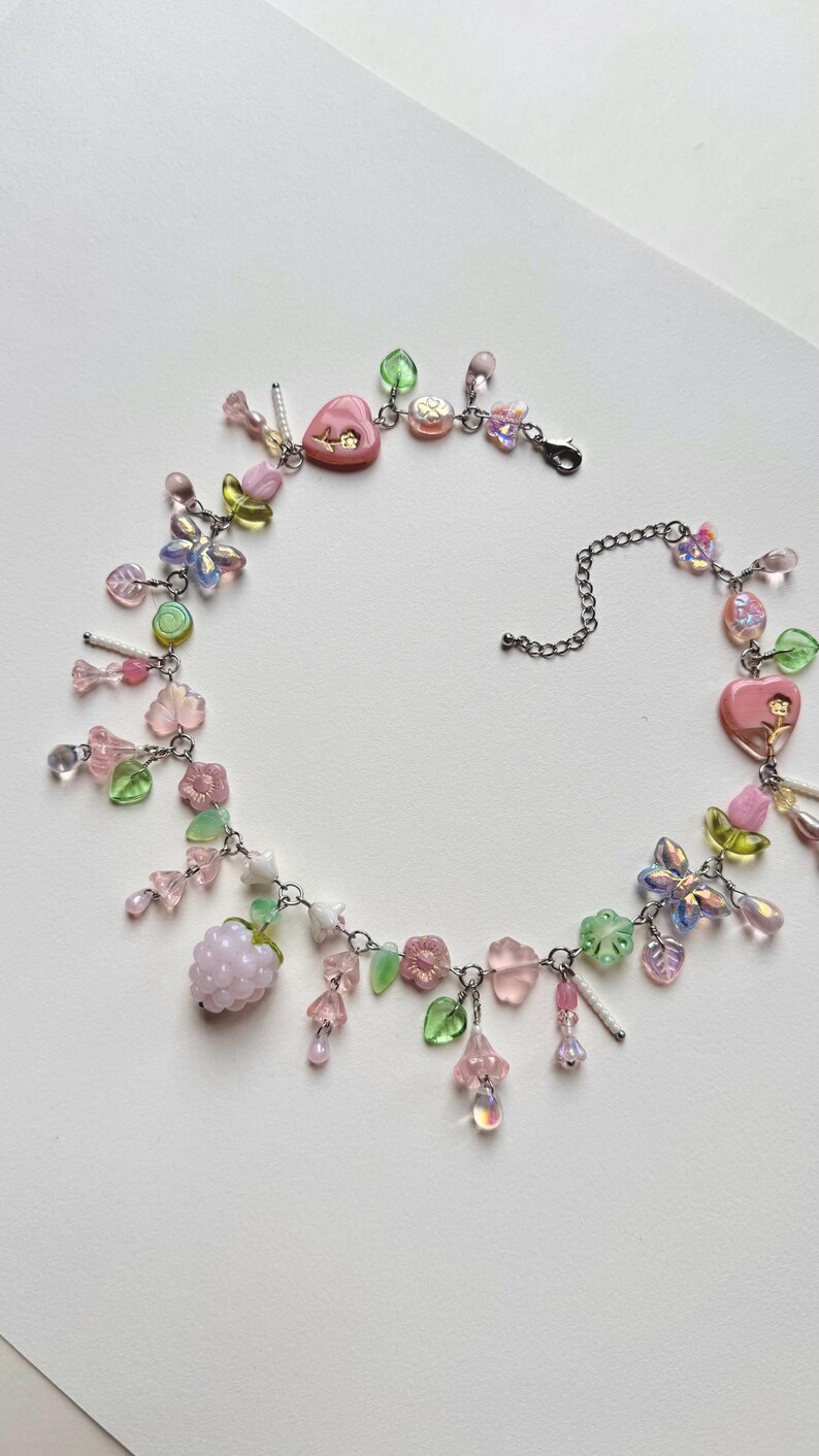 May include: A delicate necklace featuring a variety of pastel-colored beads and charms. The necklace includes pink heart-shaped pendants, flower and leaf charms, and a cluster of light purple beads. The chain is silver-toned, and the overall design is whimsical.