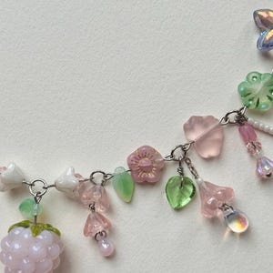 May include: A delicate necklace featuring a silver chain adorned with pink, green, and clear beads. The design includes flower and leaf charms, along with a light pink raspberry-shaped bead. The necklace is a charming accessory.