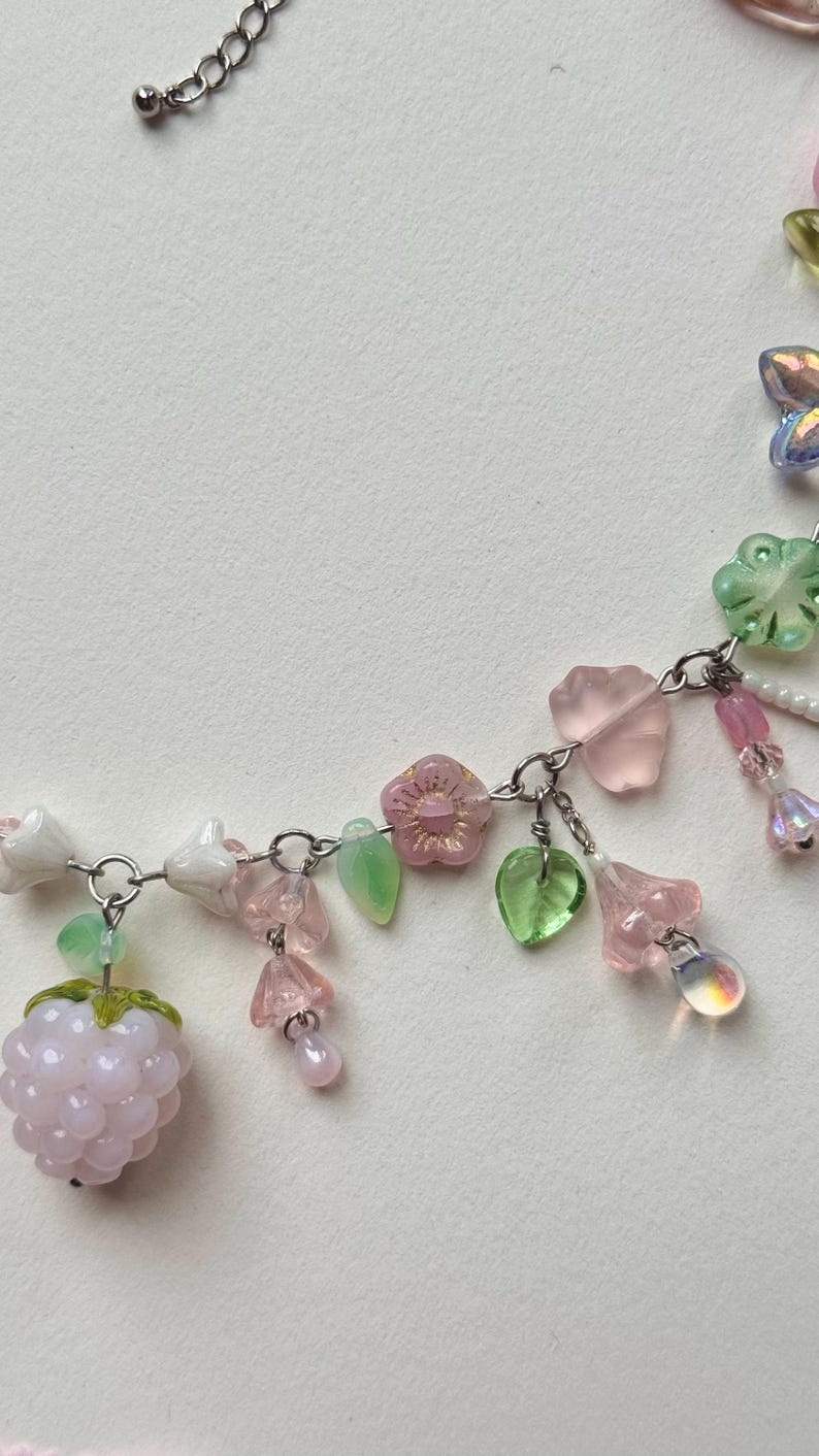 May include: A delicate necklace featuring a silver chain adorned with pink, green, and clear beads. The design includes flower and leaf charms, along with a light pink raspberry-shaped bead. The necklace is a charming accessory.