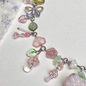 May include: A delicate necklace featuring a variety of pastel-colored beads and charms. The necklace includes pink heart-shaped beads, iridescent butterfly charms, and a cluster of pink beads resembling a raspberry. The necklace is made of metal.