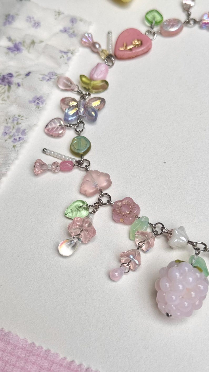 May include: A delicate necklace featuring a variety of pastel-colored beads and charms. The necklace includes pink heart-shaped beads, iridescent butterfly charms, and a cluster of pink beads resembling a raspberry. The necklace is made of metal.